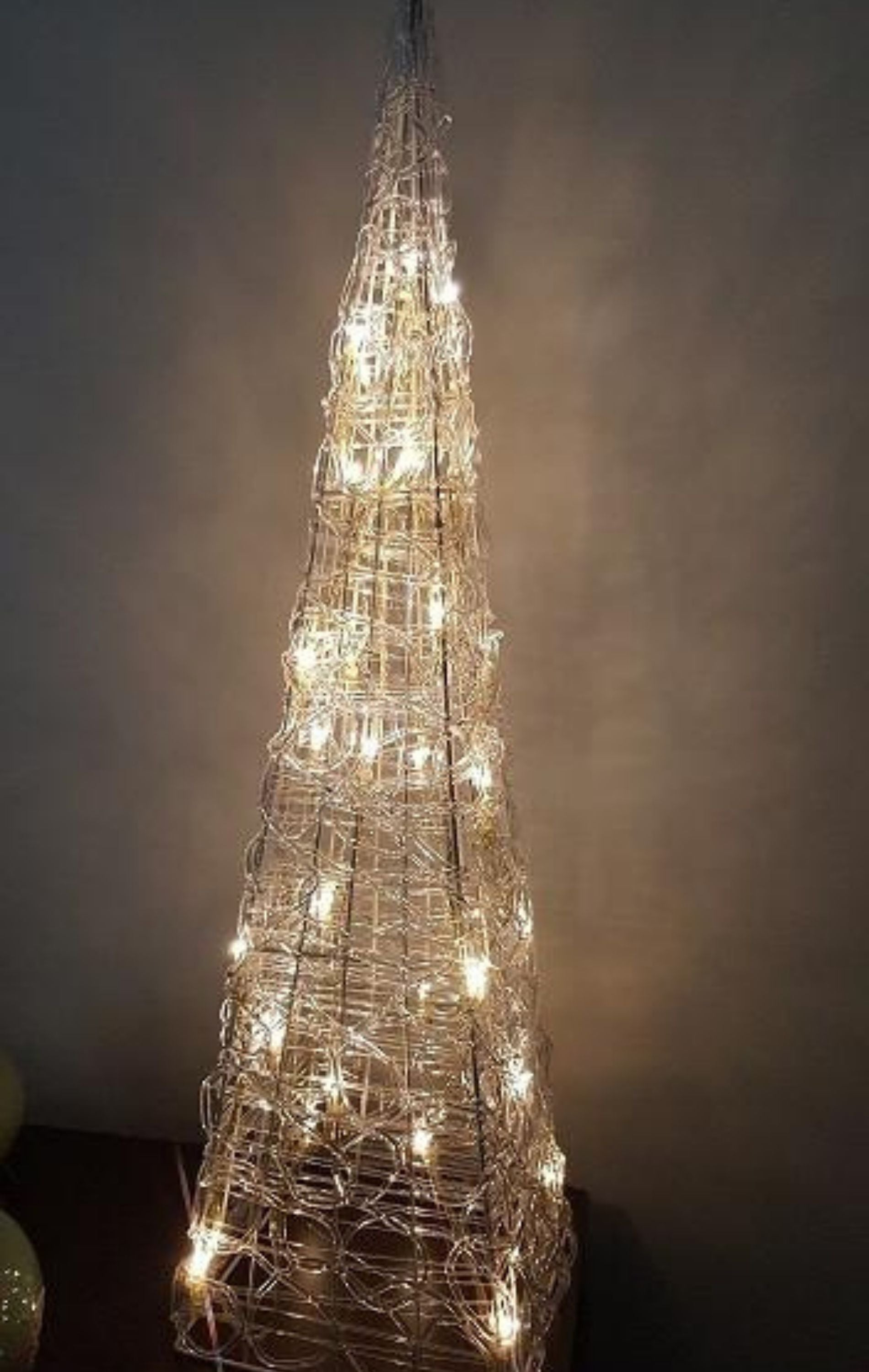 Un marco de alambre alto y brillante con forma de árbol de navidad se encuentra en el interior contra una pared lisa.