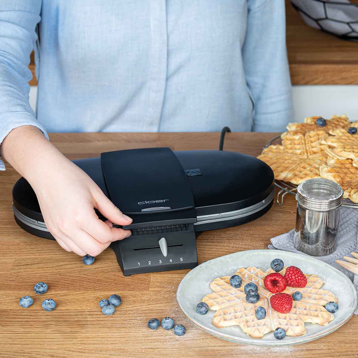 Eine Frau in blauem Hemd bedient ein Waffeleisen auf einem Holztisch mit Waffeln, Beeren und Puderzucker.