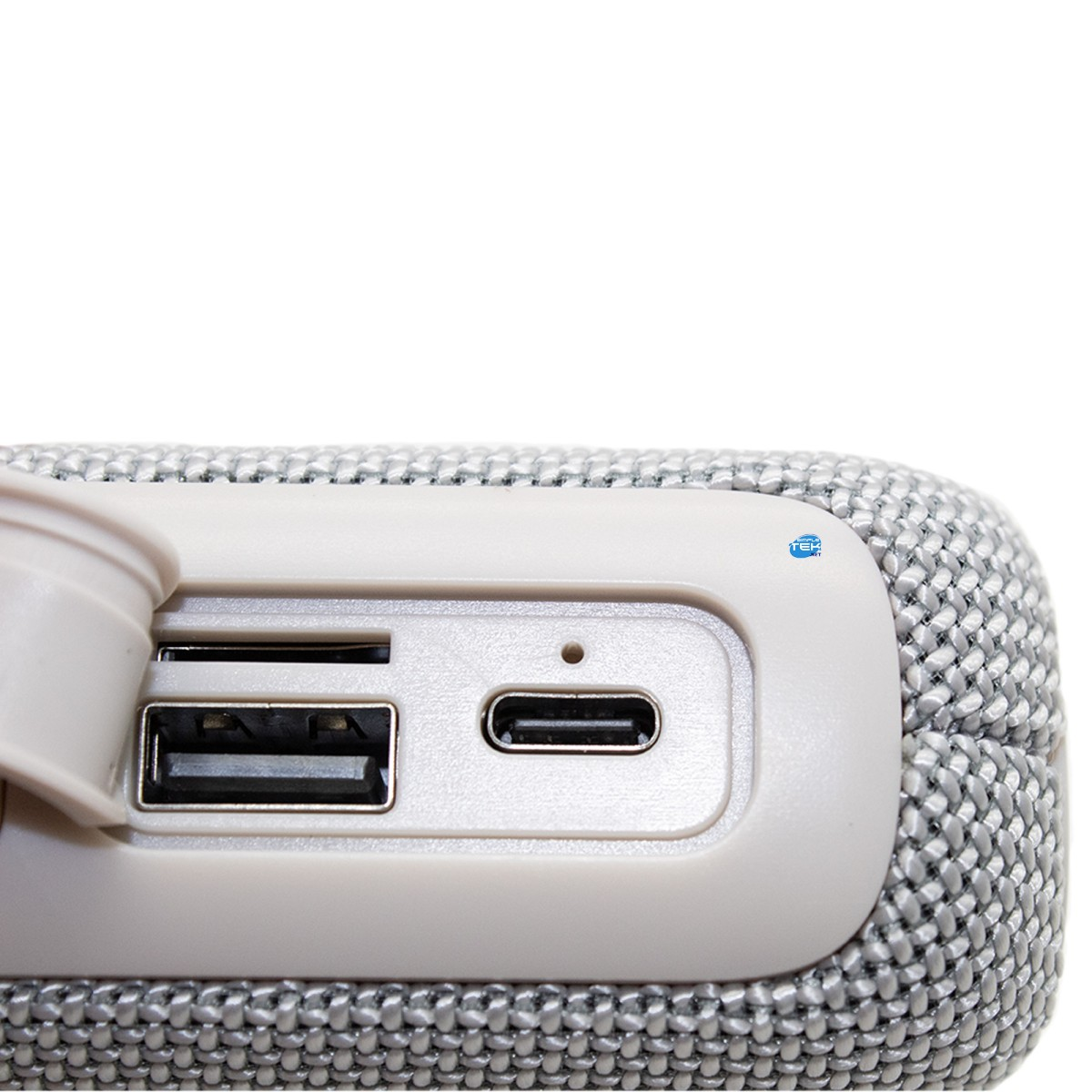 Primo piano di un altoparlante portatile in tessuto grigio con porte USB, USB-C e coperchio in gomma aperto, su fondo bianco.
