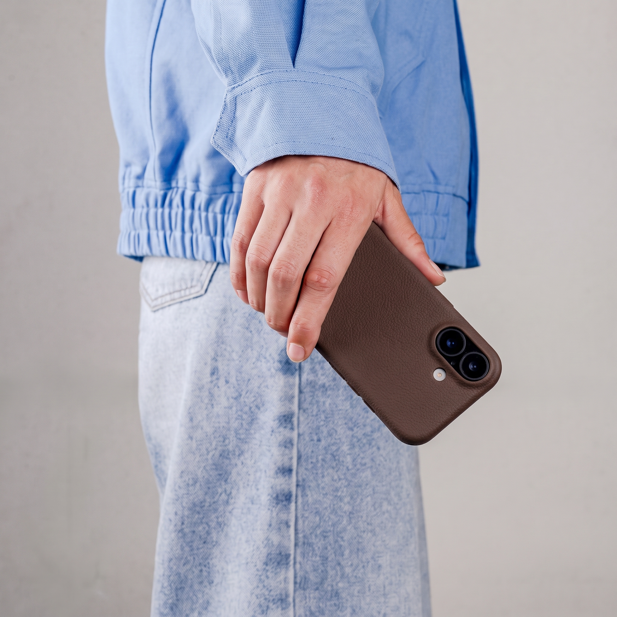 Persoon in lichtblauwe jas en jeans houdt een bruine smartphone met twee camera's vast tegen lichte achtergrond.