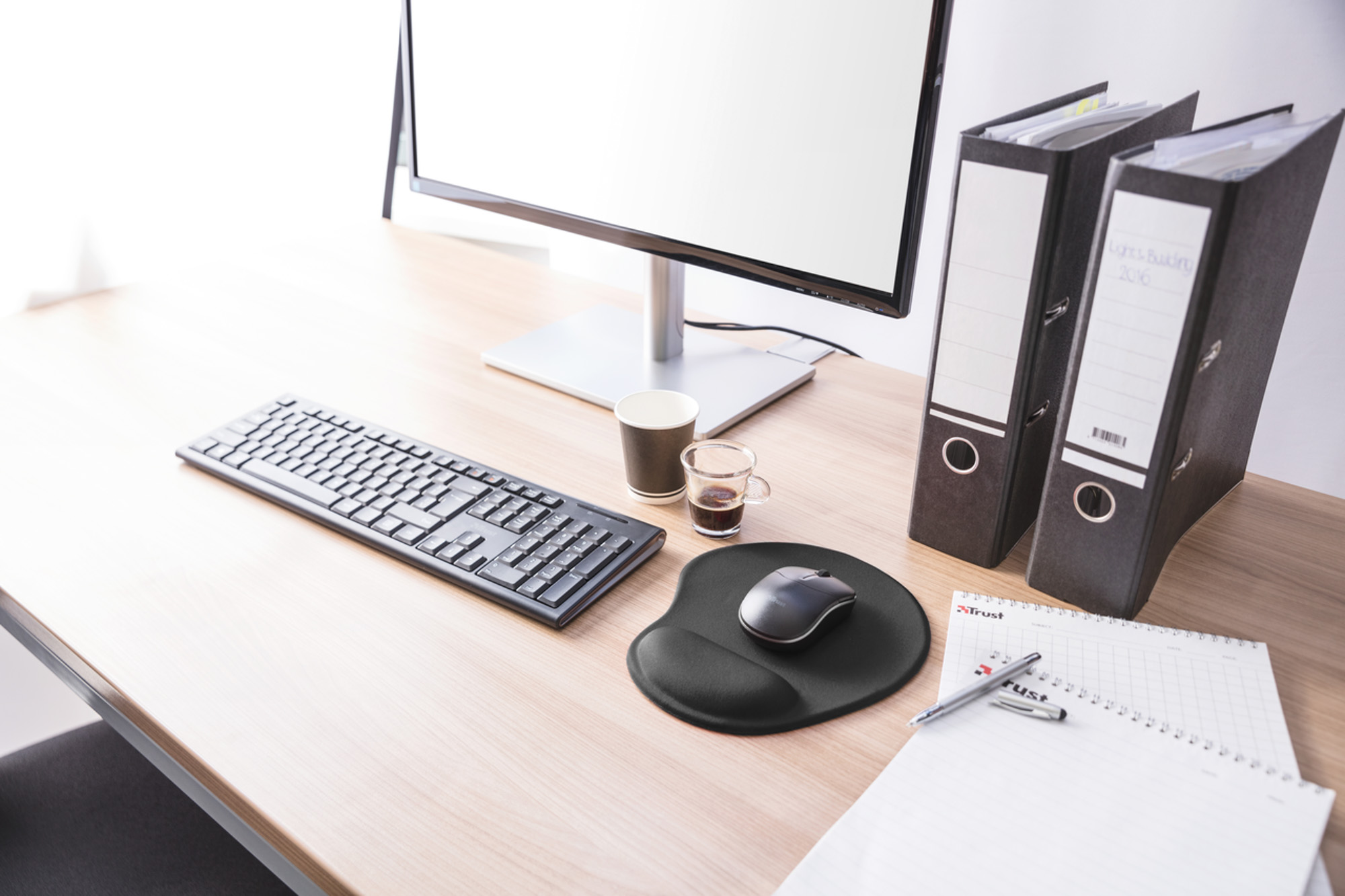 Un escritorio de madera claro con monitor, teclado, alfombrilla, tazas de café, archivadores y bloc con bolígrafo.