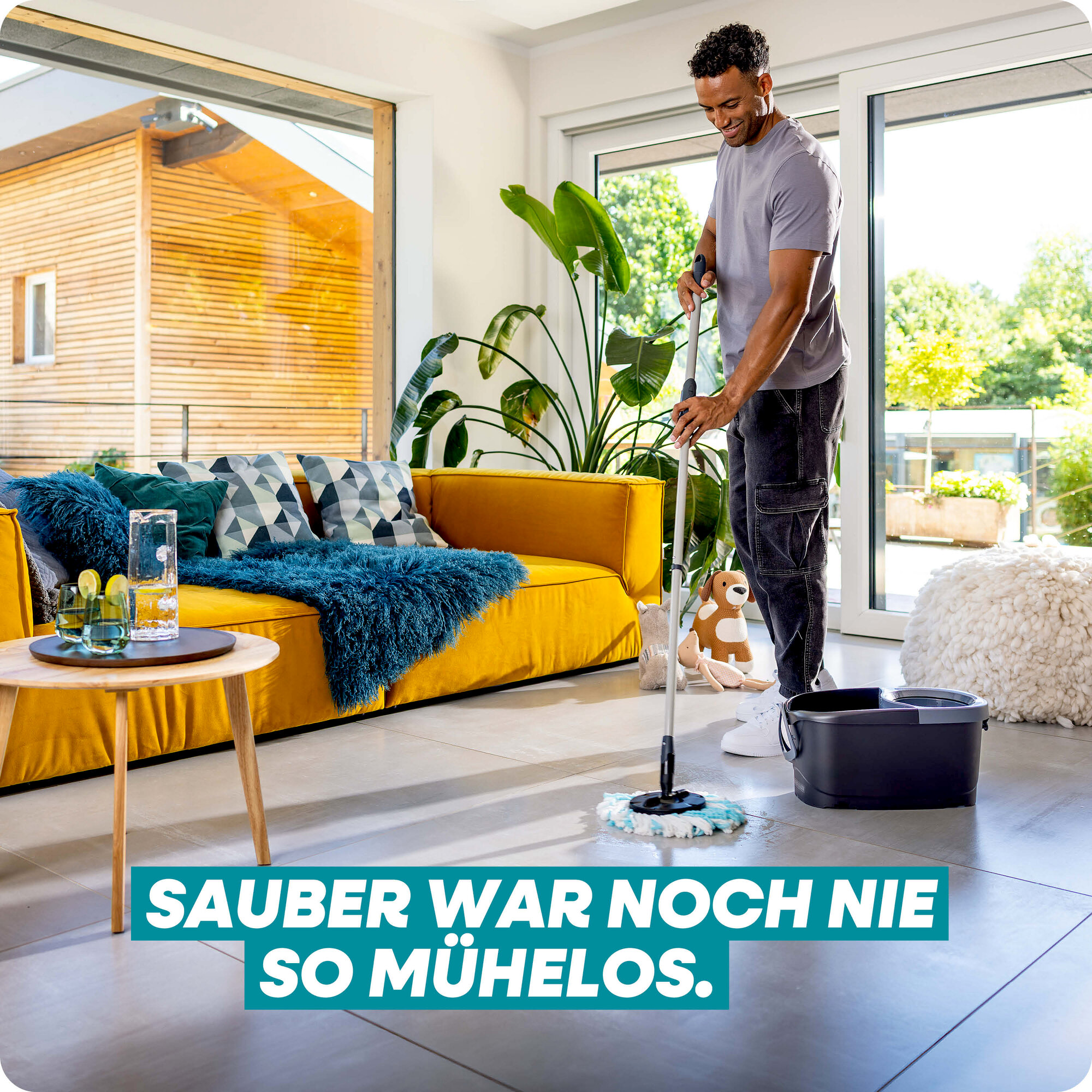 Ein Mann wischt den Boden eines hellen Wohnzimmers neben einem gelben Sofa.