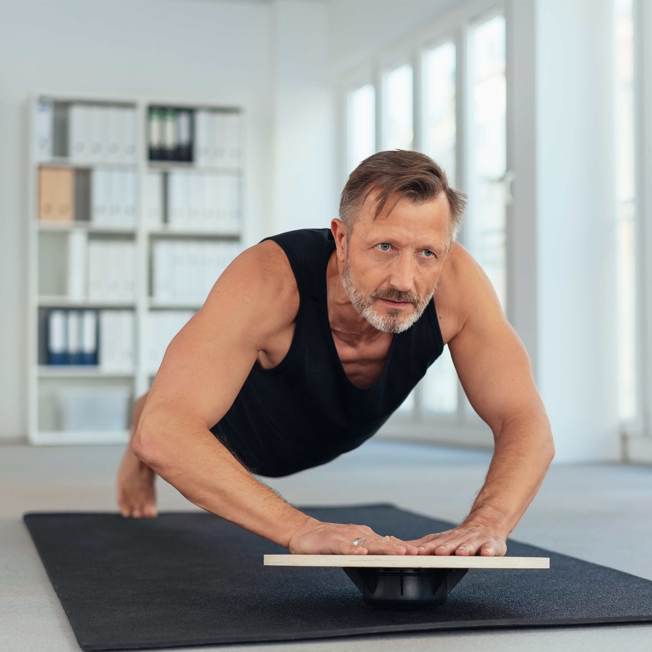 Ein reifer Mann in schwarzem Tanktop macht Liegestütze auf einem Balance Board und einer Fitnessmatte im Büro.
