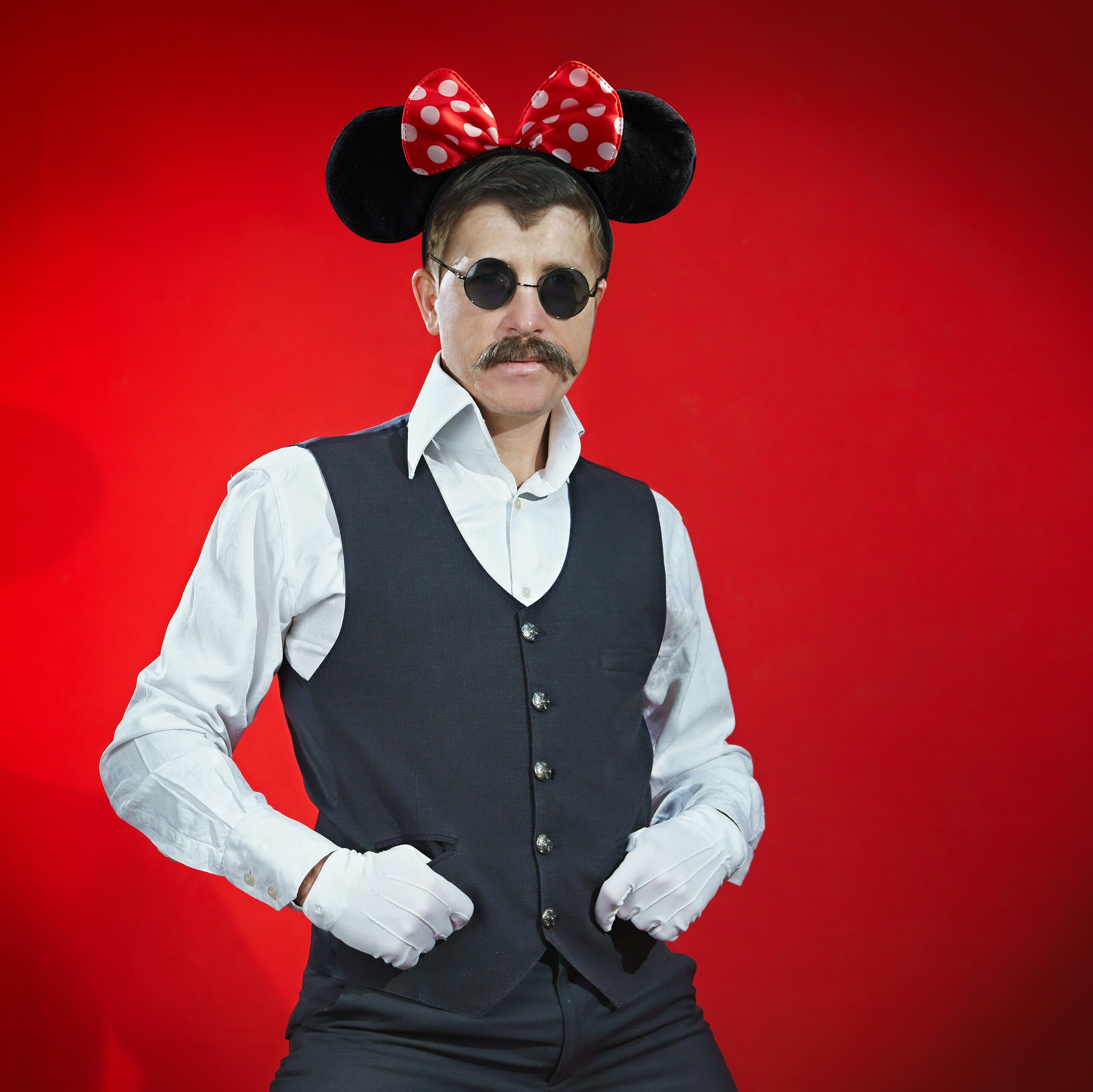 Ein Mann mit Schnurrbart, Sonnenbrille und Minnie-Maus-Ohren steht vor rotem Hintergrund.