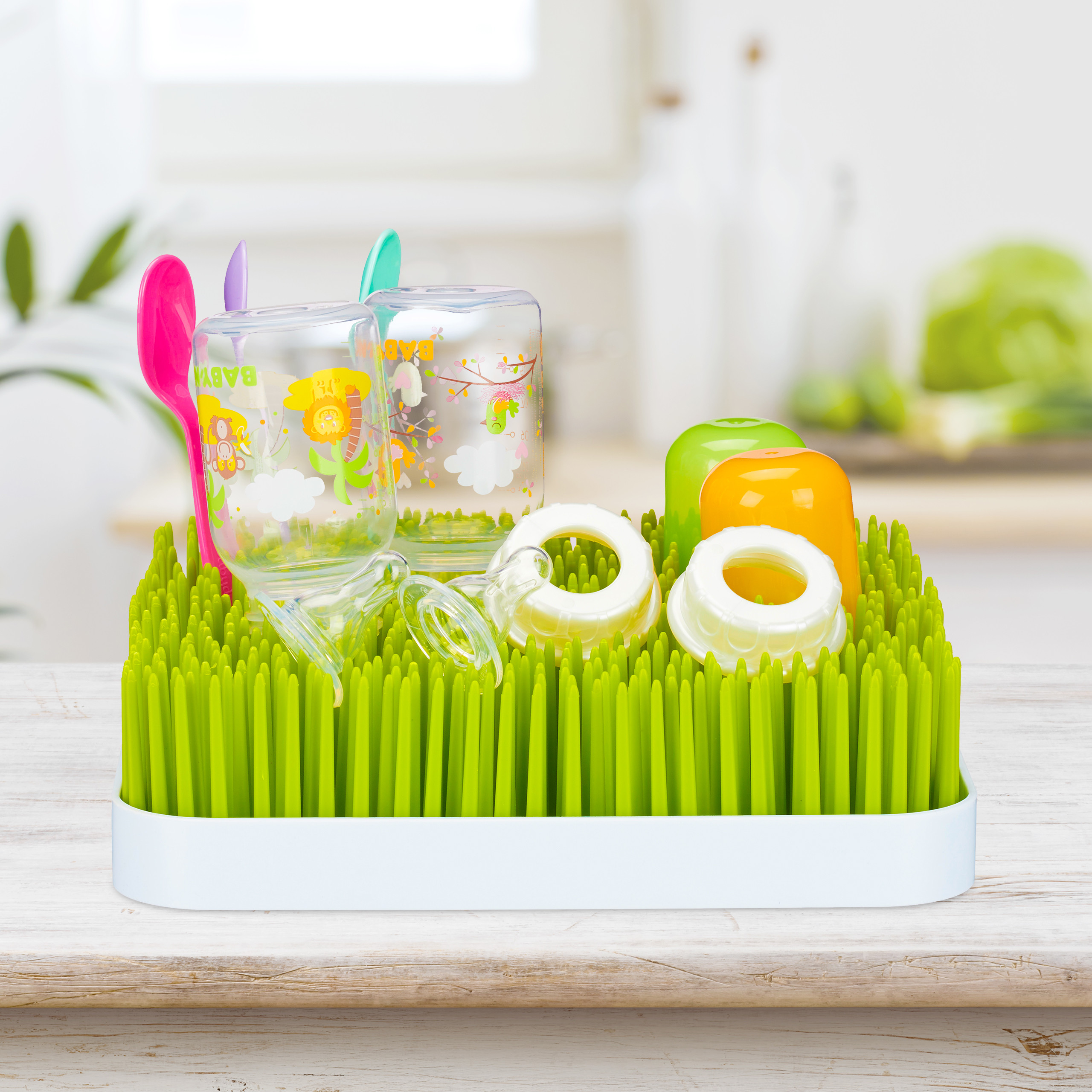 Ein grünes grasähnliches Babydunstgestell auf einem Holztisch hält Flaschen, Sauger und bunte Löffel.