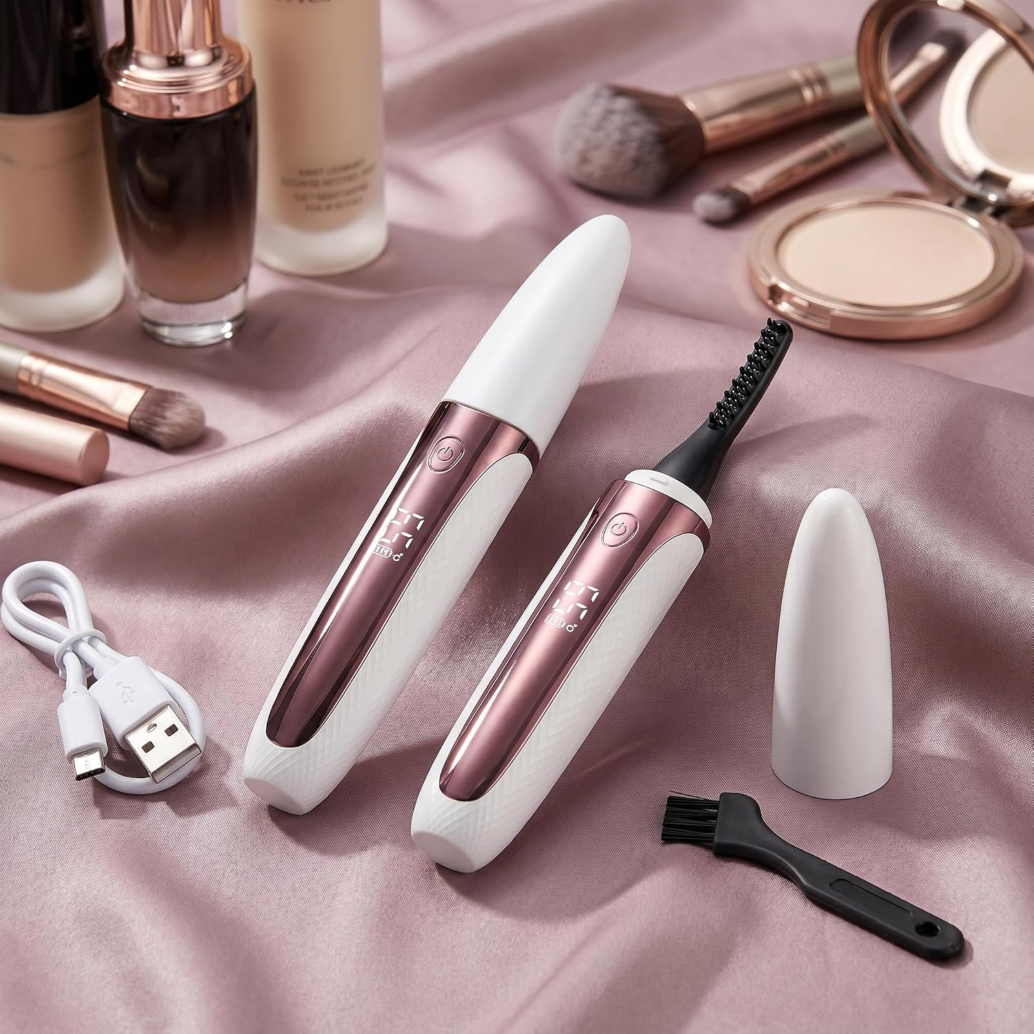 Zwei rosévergoldete und weiße beheizbare Wimpernzangen liegen auf einem rosa Satintuch neben Make-up-Artikeln.