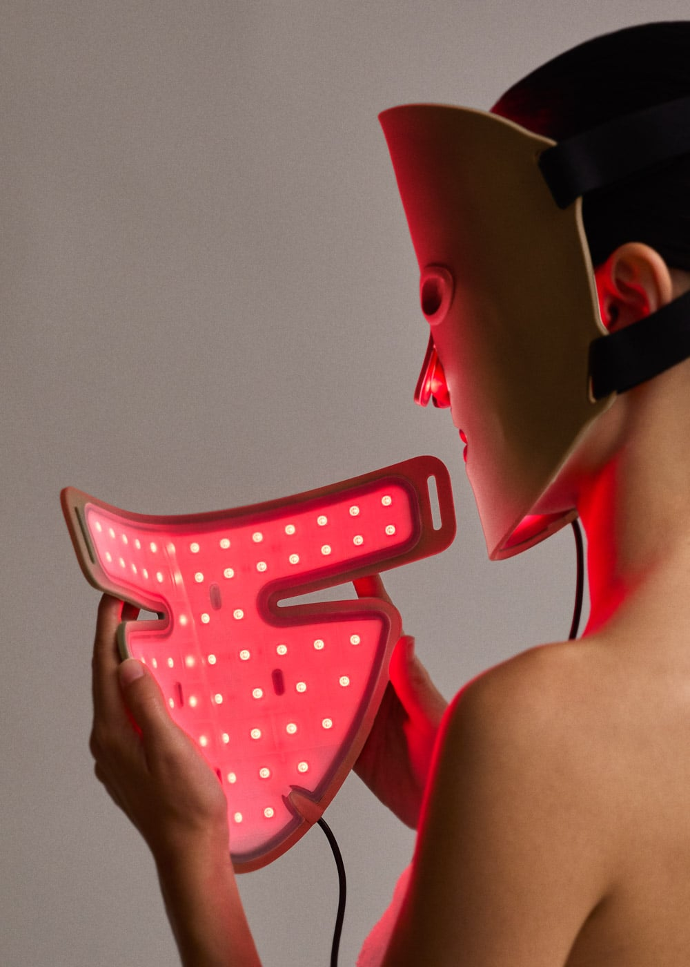 Una persona lleva una mascarilla facial LED beige y sostiene una mascarilla para el cuello roja brillante.