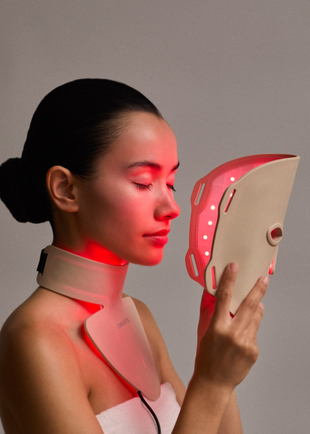 Una mujer con los ojos cerrados sostiene una máscara facial LED roja y lleva un collarín a juego.