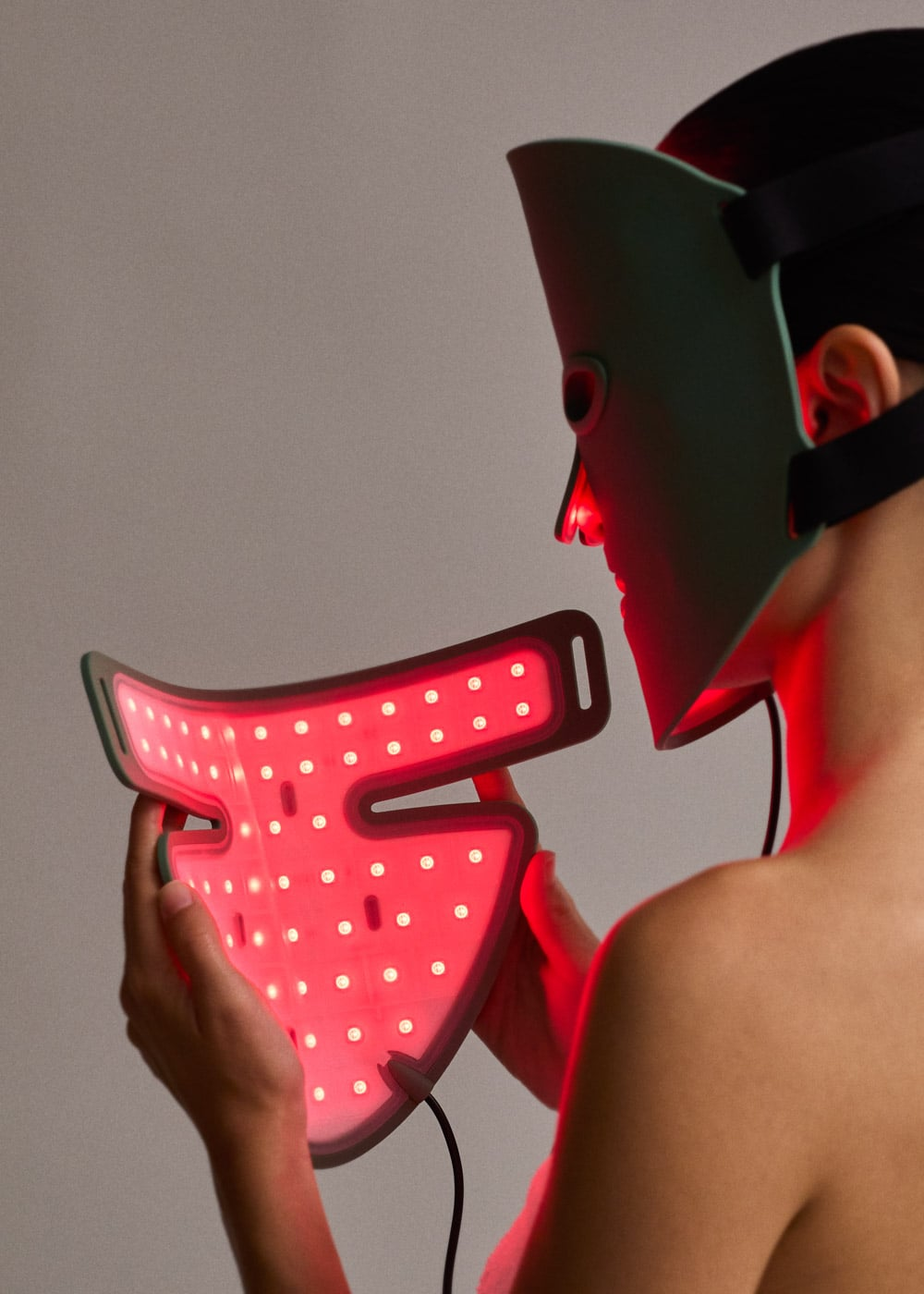Una persona con una mascarilla facial LED verde sostiene otra mascarilla de terapia de luz roja brillante.