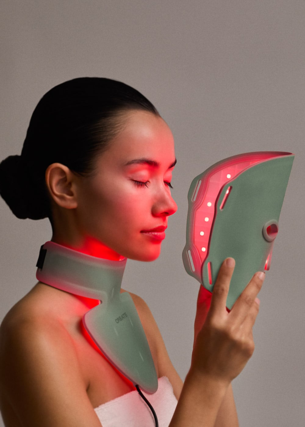 Una mujer sostiene una mascarilla facial LED verde y lleva un collar a juego con luz roja sobre fondo gris.