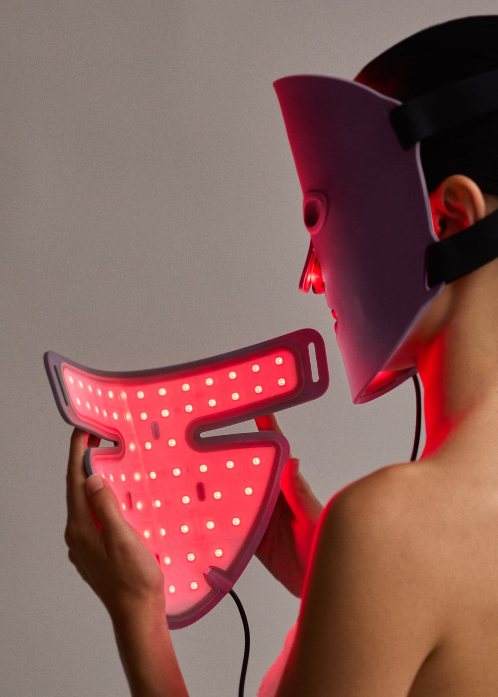 Una persona lleva una mascarilla facial LED morada y sostiene una segunda mascarilla de luz roja brillante.