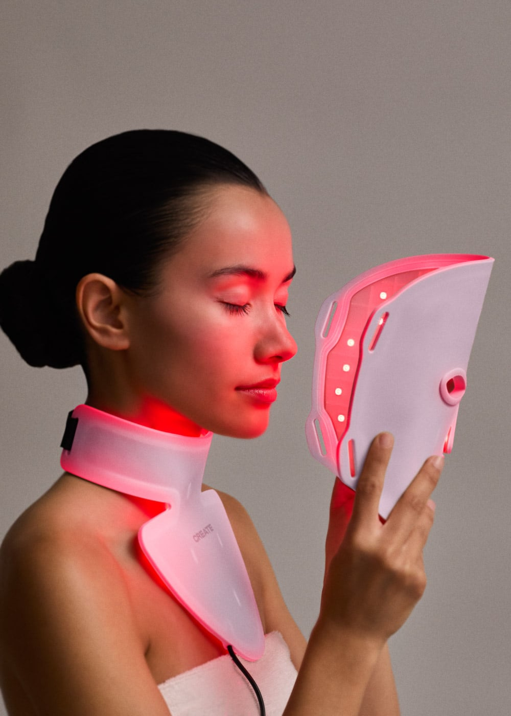 Una mujer sostiene una mascarilla facial LED blanca con luz roja, una pieza para el cuello brilla en rojo.