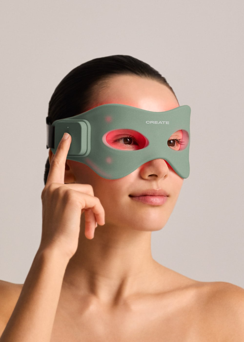 Una mujer con una máscara ocular de terapia de luz verde, tocando el botón de control en el lateral sobre fondo neutro.