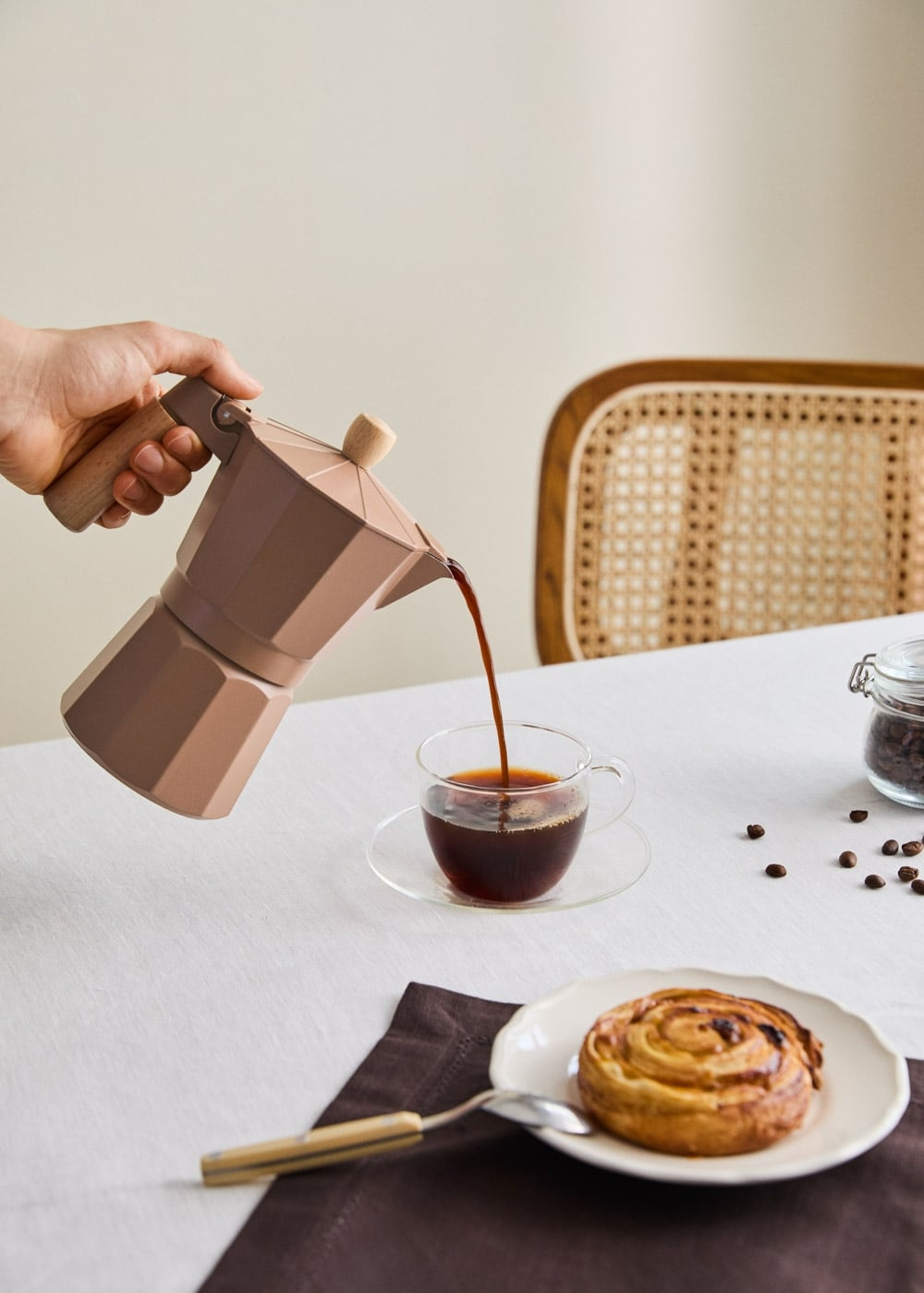Una mano vierte café de una cafetera marrón en una taza sobre una mesa puesta con bollería.