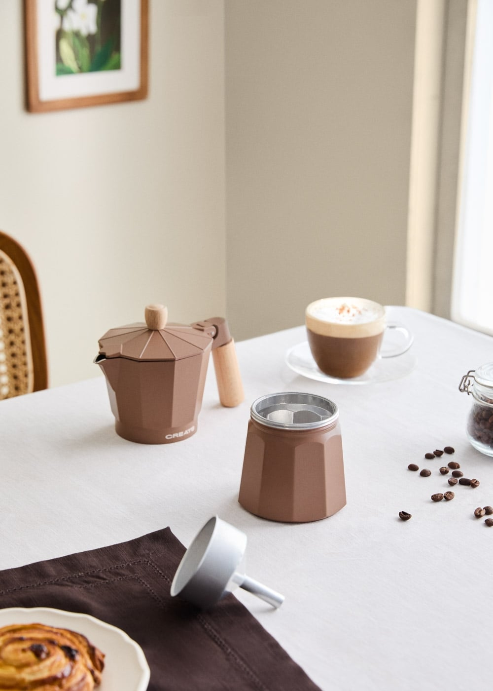Una cafetera moka marrón, granos de café, pastelería y un capuchino sobre un mantel blanco.