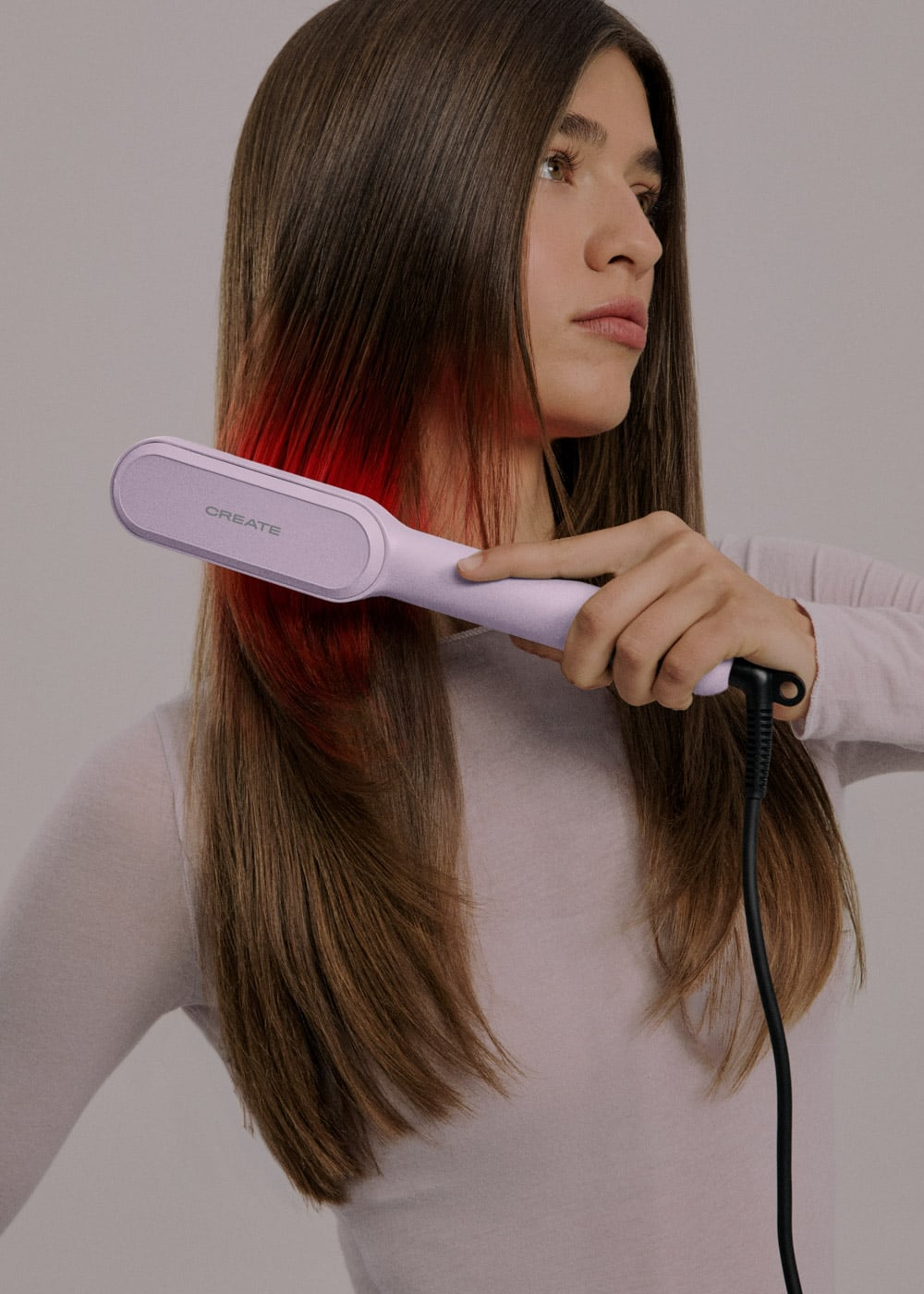 Una mujer usa una plancha de pelo lila claro, peinando su largo cabello castaño sobre un fondo gris.