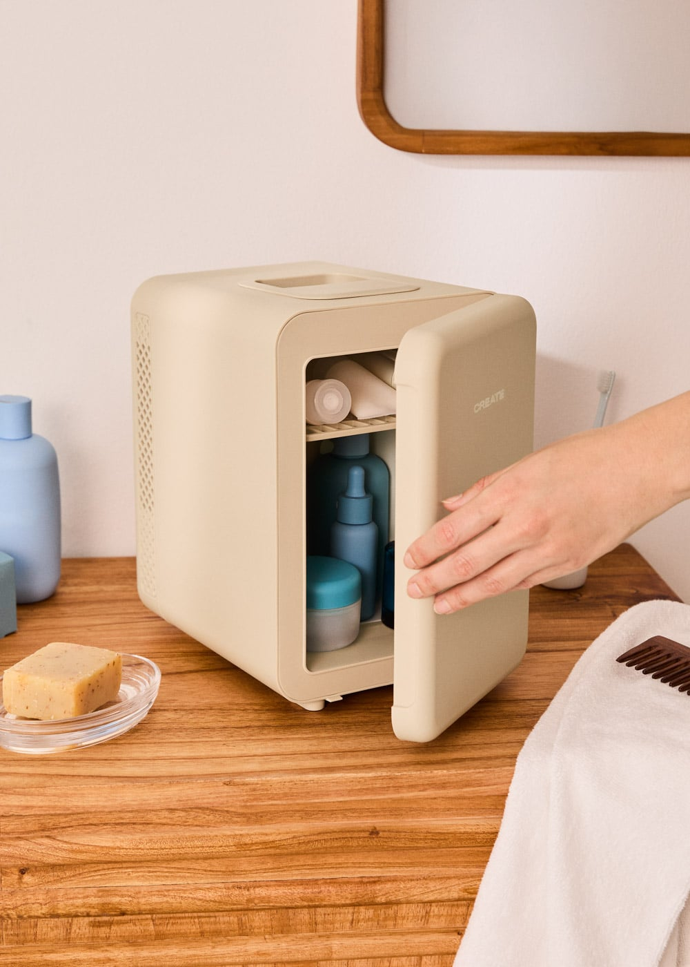 Una mano abre una pequeña nevera de cosméticos beige en una mesa de madera con productos.