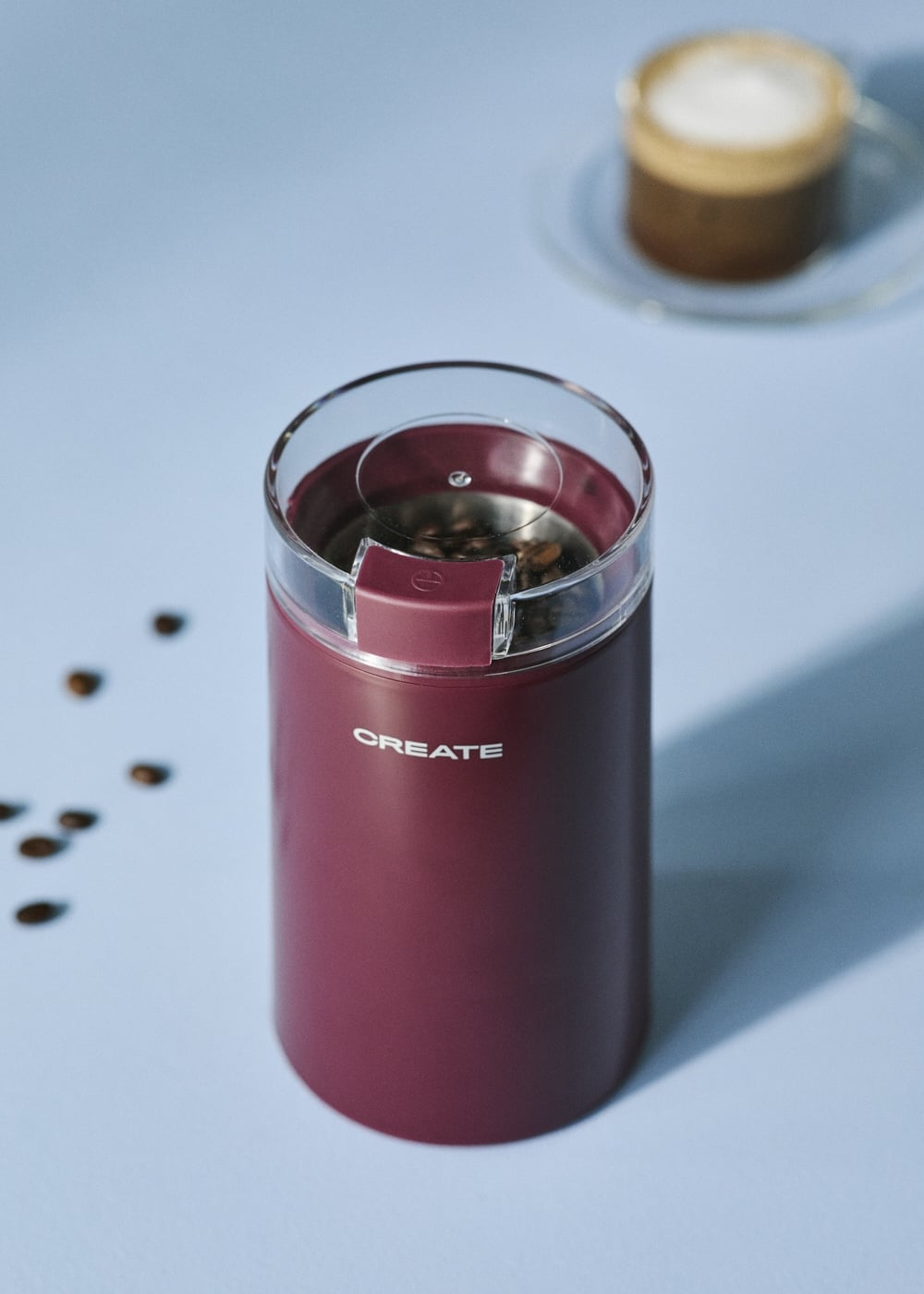 Un molinillo de café eléctrico burdeos sobre fondo azul claro con granos de café y taza de café al fondo.