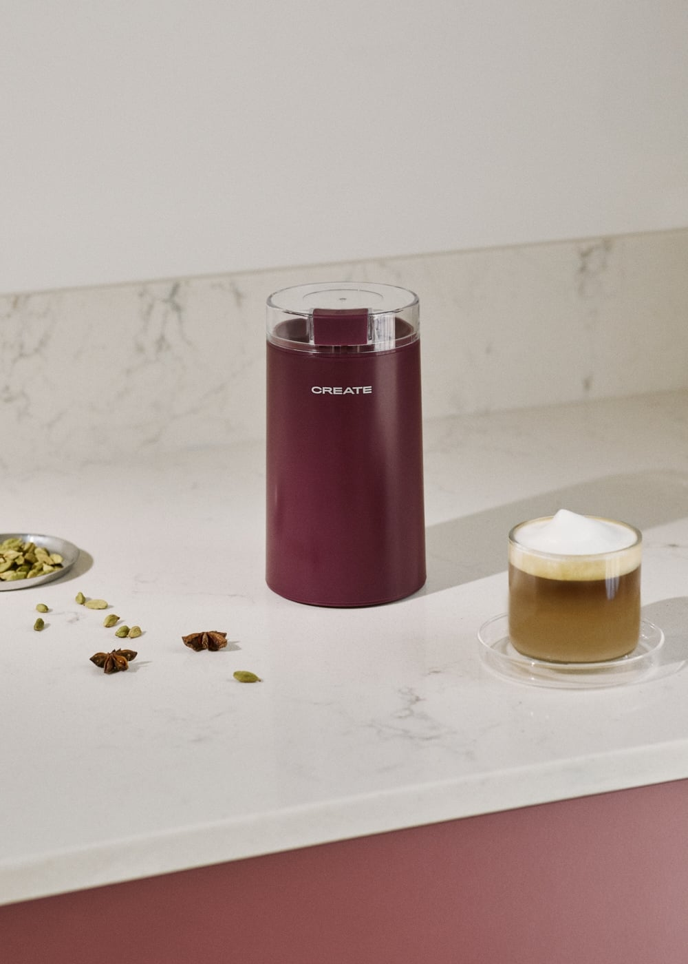 Un molinillo de café granate en una encimera de mármol blanco, con anís estrellado, cardamomo y un capuchino.
