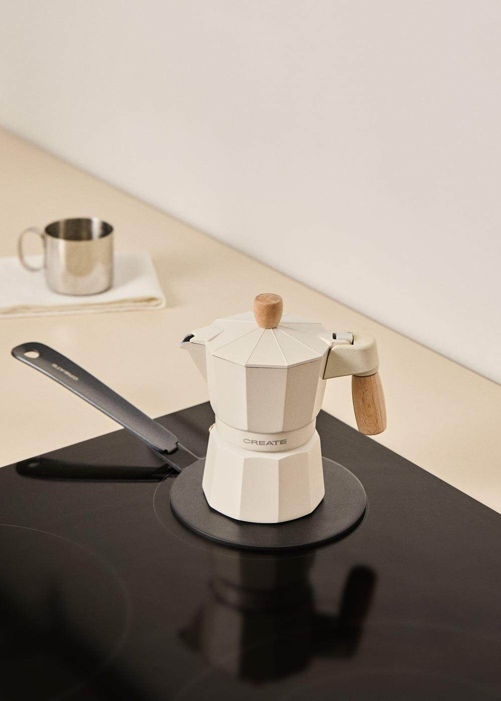 Una cafetera italiana blanca con asas de madera sobre una placa de inducción negra, con una taza plateada al fondo.
