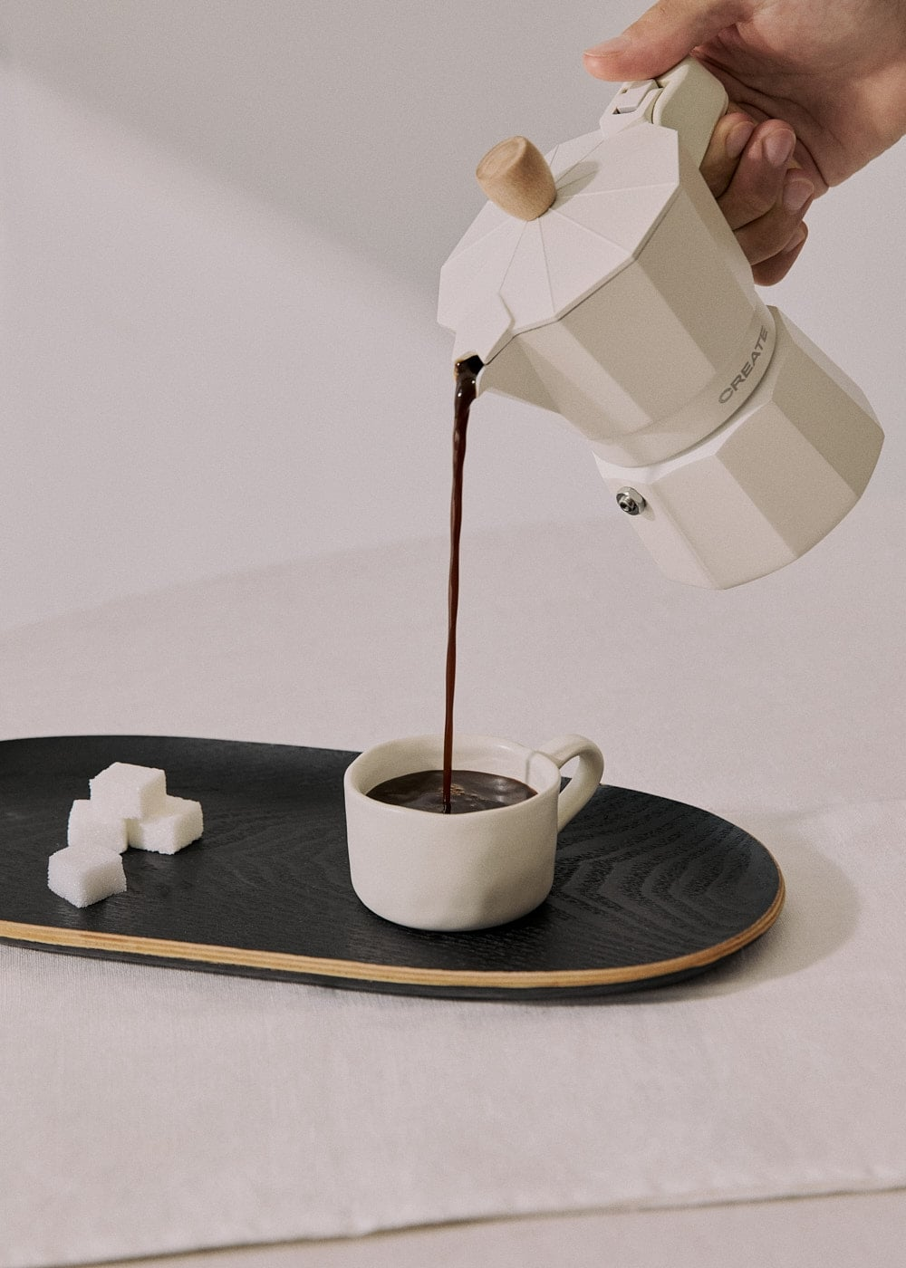 Una mano vierte café de una cafetera moka blanca en una taza blanca sobre una bandeja negra con terrones de azúcar.