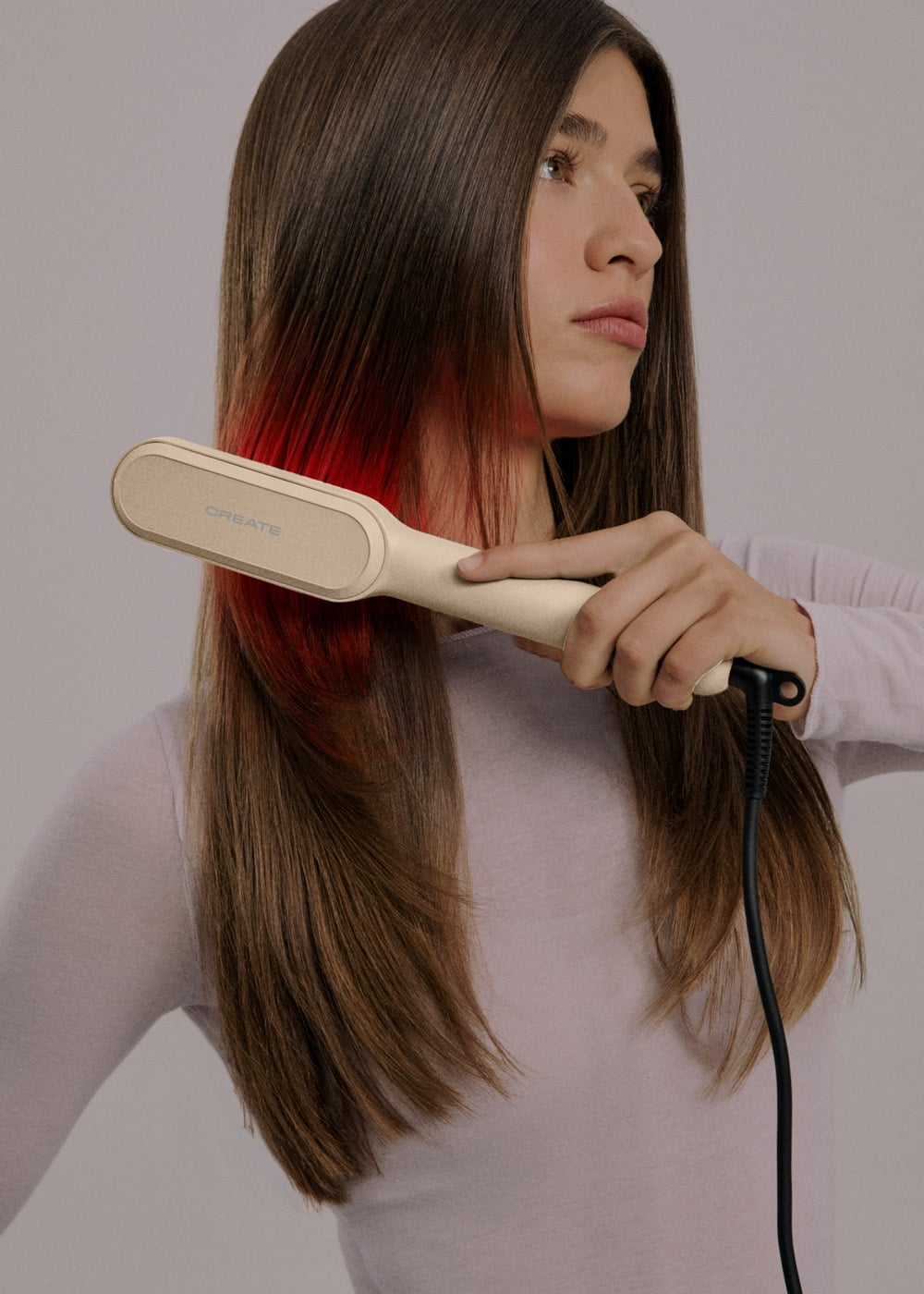 Una mujer con top blanco alisa su pelo largo castaño con una plancha de pelo beis que brilla en rojo.