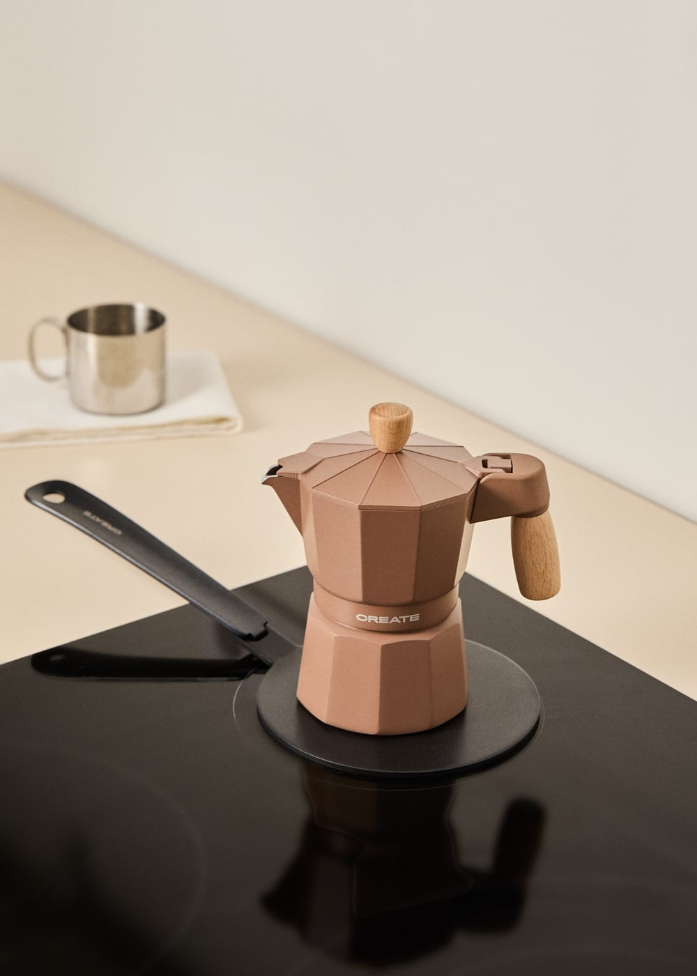 Una cafetera moka de oro rosa con asas de madera sobre una placa de inducción negra; al fondo, una taza metálica.