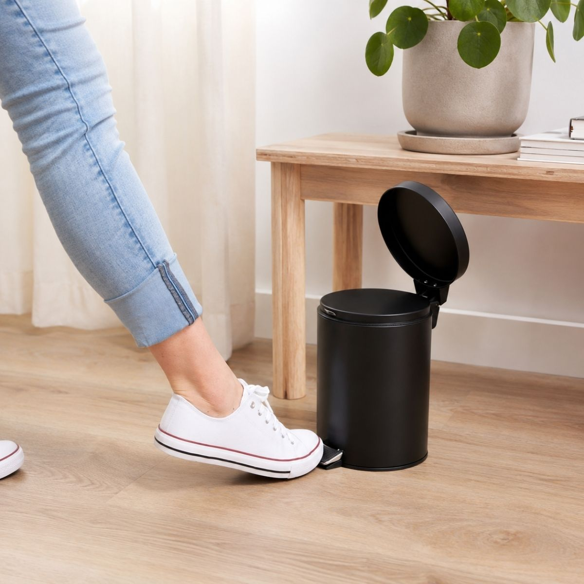 A person in jeans and white sneakers steps on the pedal of a black trash can next to a wooden table.