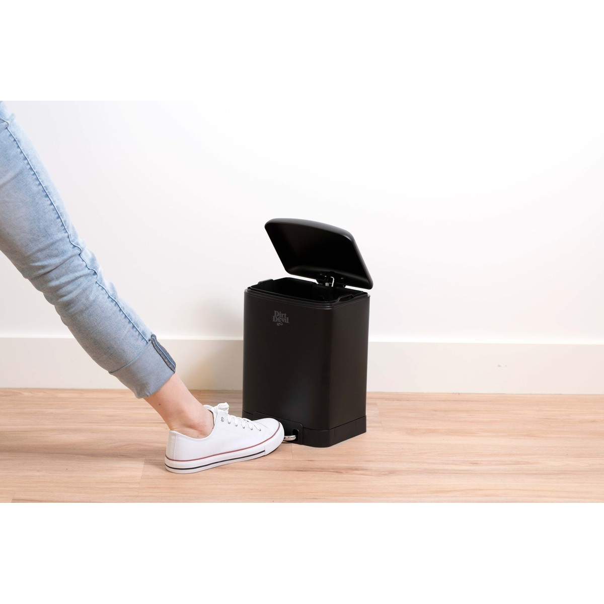 Une personne en jean et baskets blanches appuie sur la pédale d'une poubelle noire sur un sol en bois.