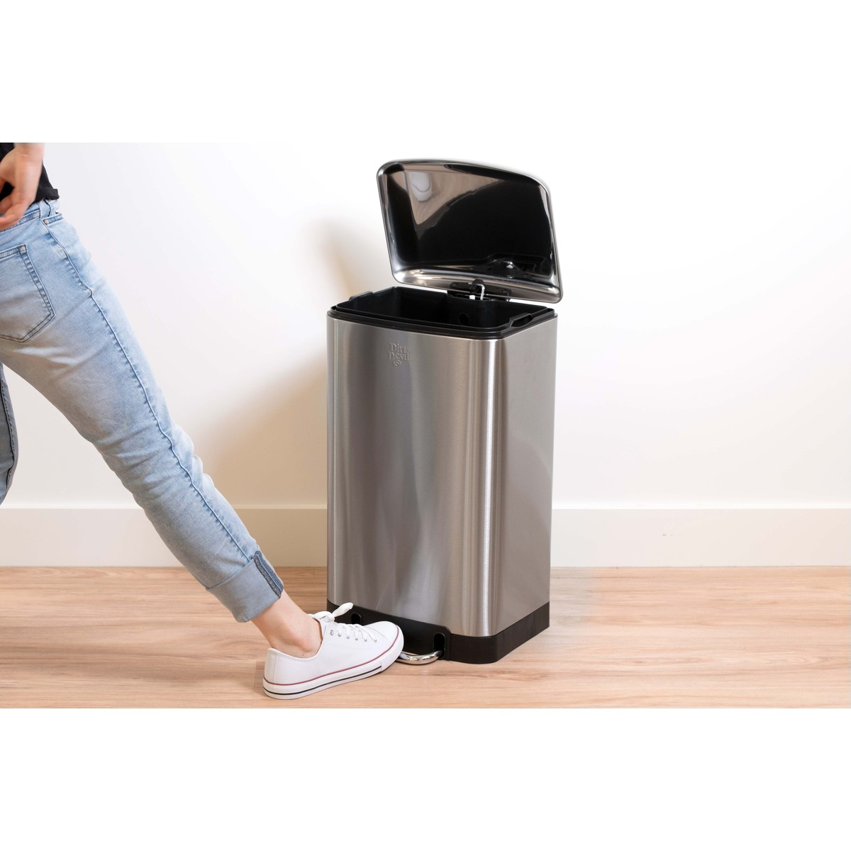 Persoon in jeans en witte sneakers trapt op het pedaal van een roestvrijstalen prullenbak met open deksel.