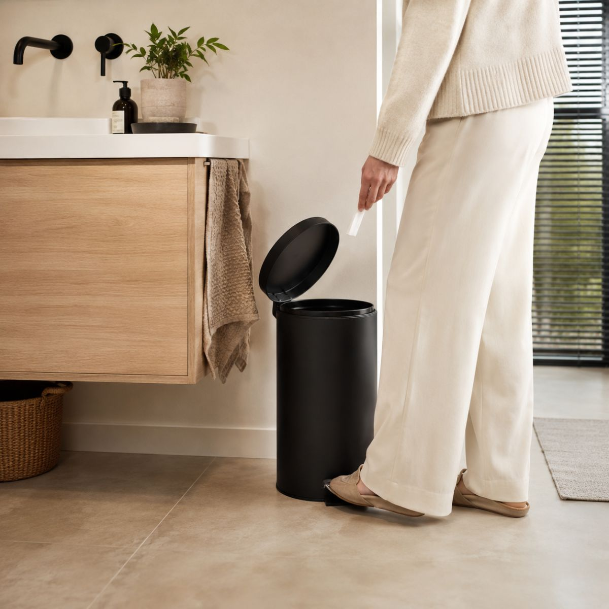 Une personne en vêtements clairs ouvre une poubelle à pédale noire dans une salle de bain.