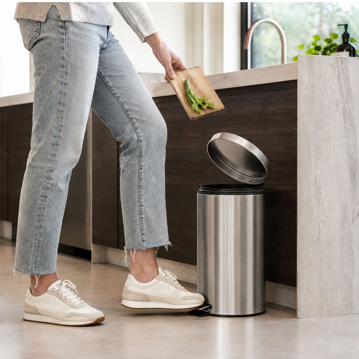 Une personne en jean et baskets blanches appuie sur une pédale pour ouvrir une poubelle de cuisine.