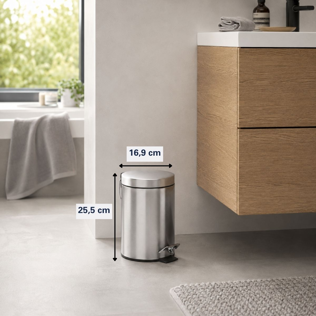 A small silver pedal bin with dimensions 16.9 cm wide and 25.5 cm tall stands in a bathroom.