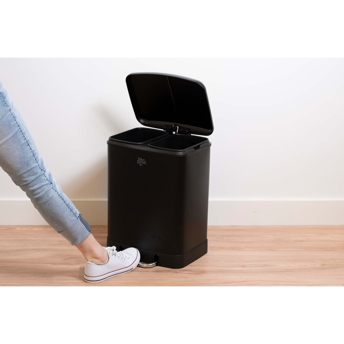 Un pied appuie sur la pédale d'une poubelle noire à deux compartiments, ouvrant son couvercle.