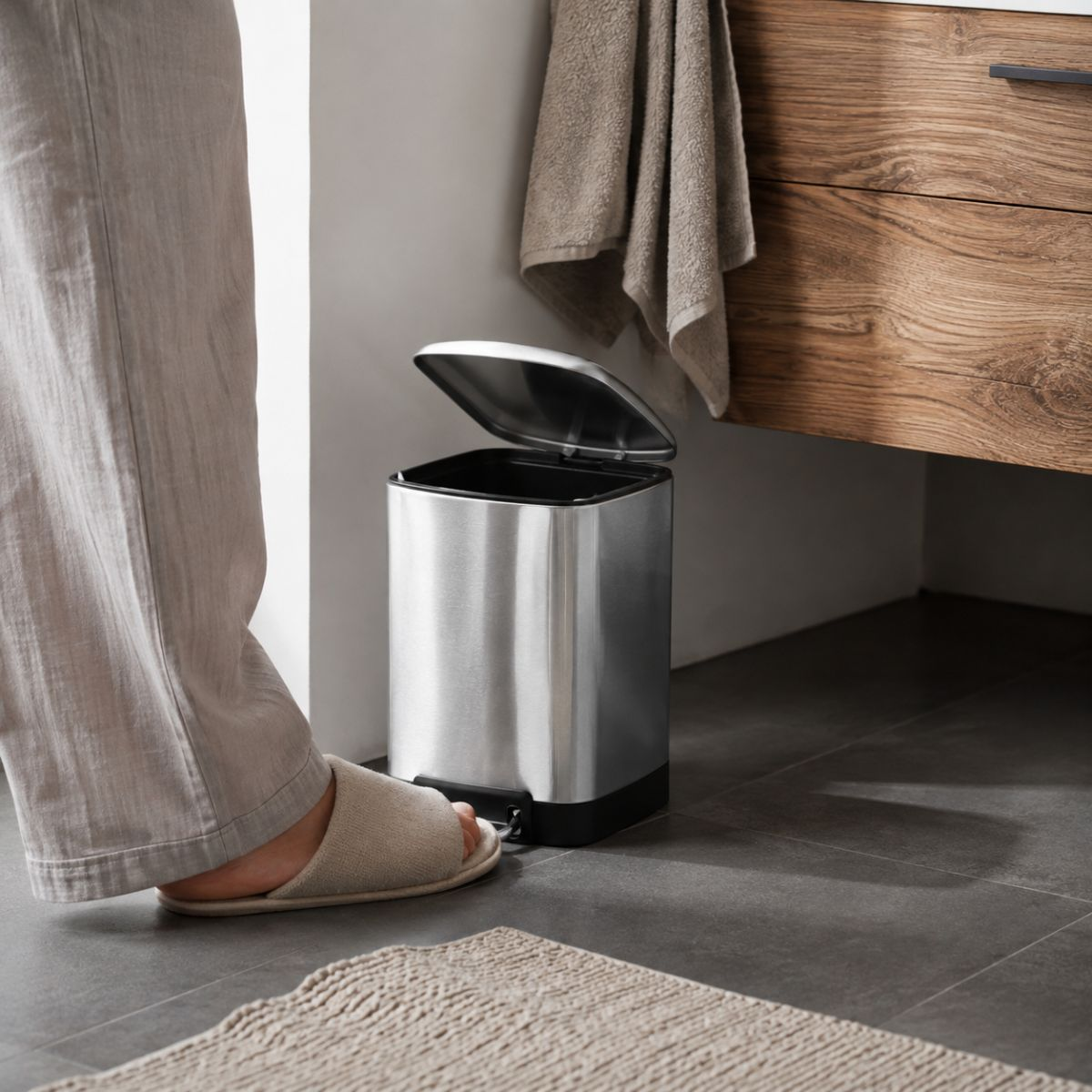 Un pied en pantoufle appuie sur la pédale d'une petite poubelle en acier inoxydable dans une salle de bain.