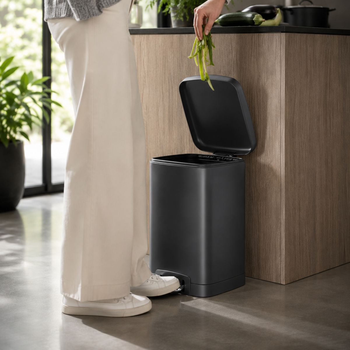 Person throwing vegetable scraps into an open black pedal bin in a modern kitchen.