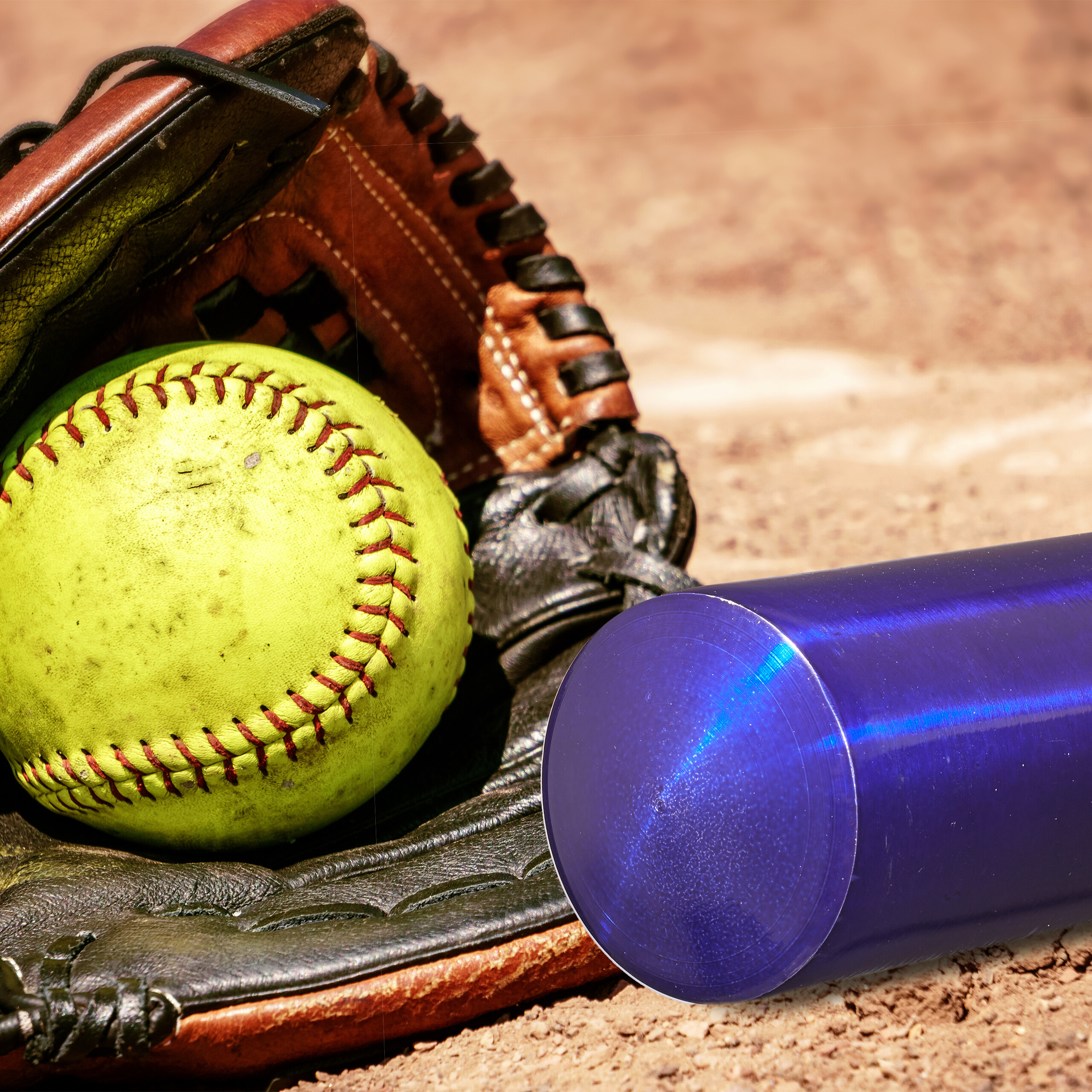Ein gelber Softball in einem braunen und schwarzen Handschuh neben einem blauen Schläger auf sandigem Boden.