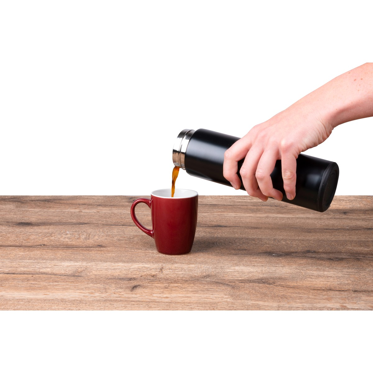 Eine Hand giesst Kaffee aus einer schwarzen Thermoskanne in eine rote Tasse auf einem Holz Tisch.