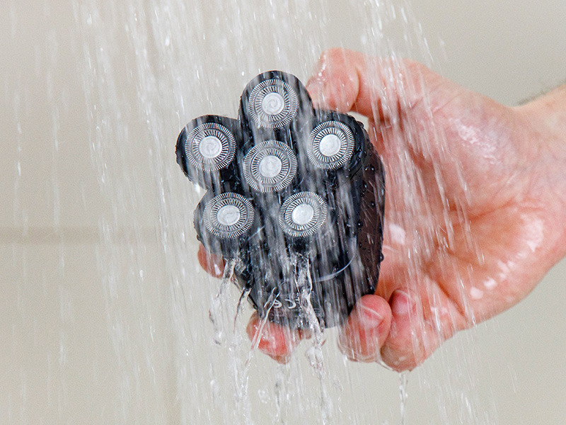 Una mano tiene un rasoio nero a cinque teste sotto l'acqua corrente della doccia.
