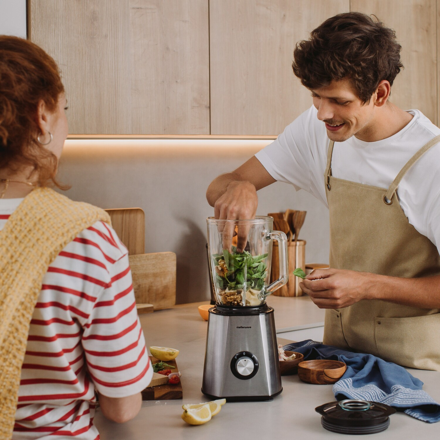 Una pareja prepara un batido en una cocina moderna, mientras el hombre añade ingredientes a la batidora.