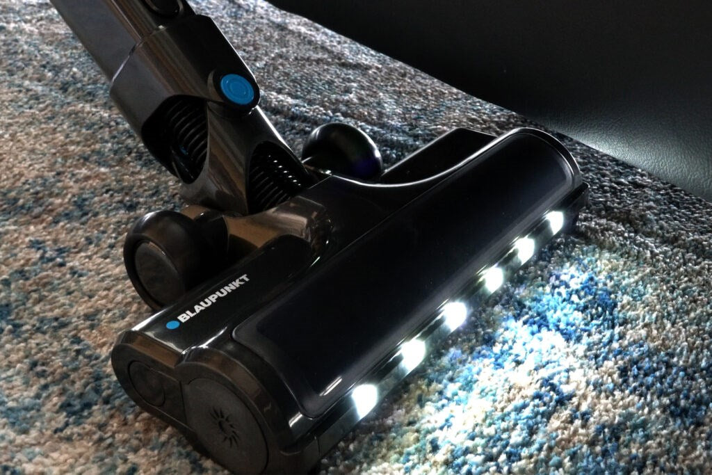 Ein schwarzer Blaupunkt Staubsaugerkopf mit leuchtenden LED-Ländern auf einem blau-weiß gemusterten Teppich.