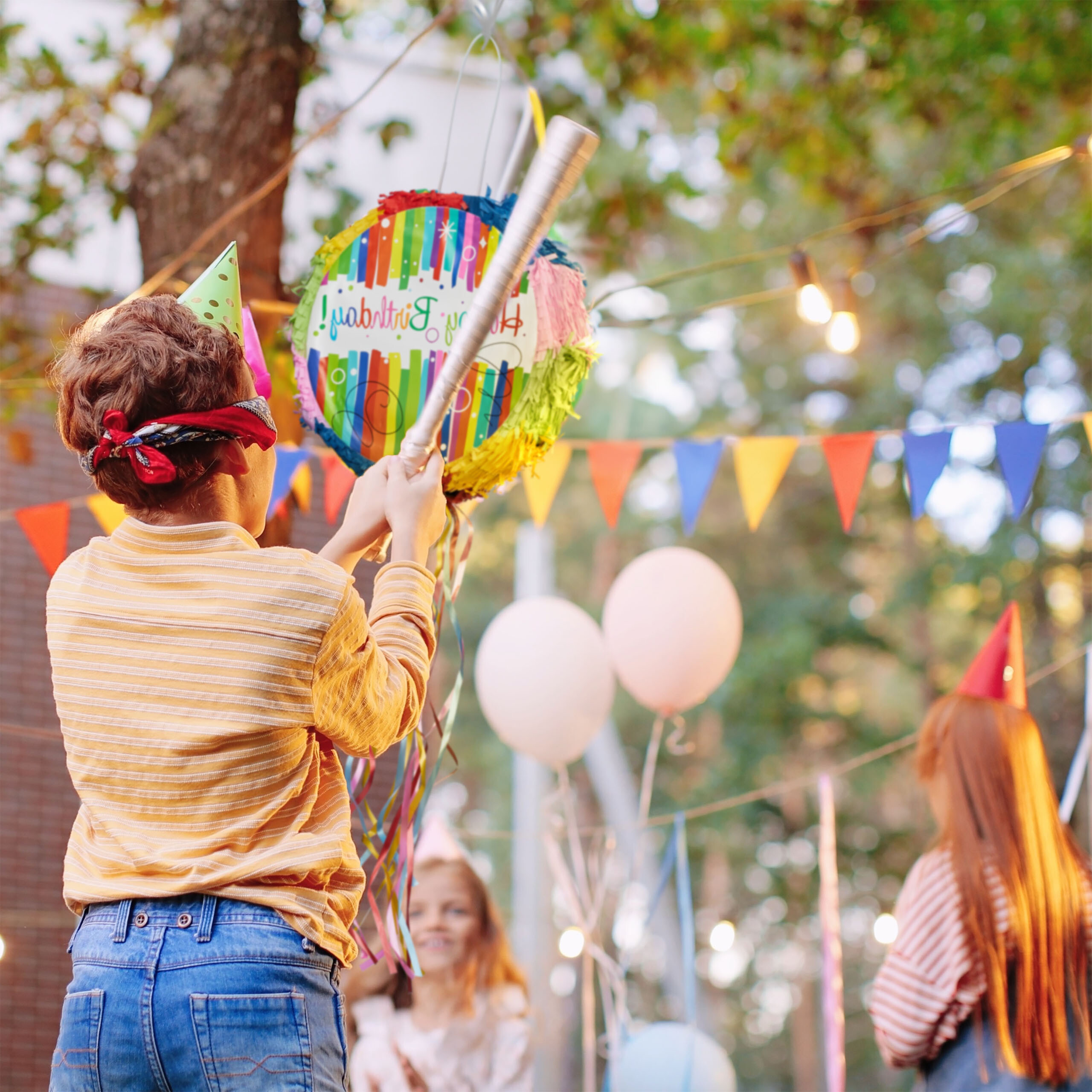 Ein Junge mit verbundenen Augen und Partyhut schlägt bei einer Geburtstagsfeier im Freien auf eine bunte Piñata.