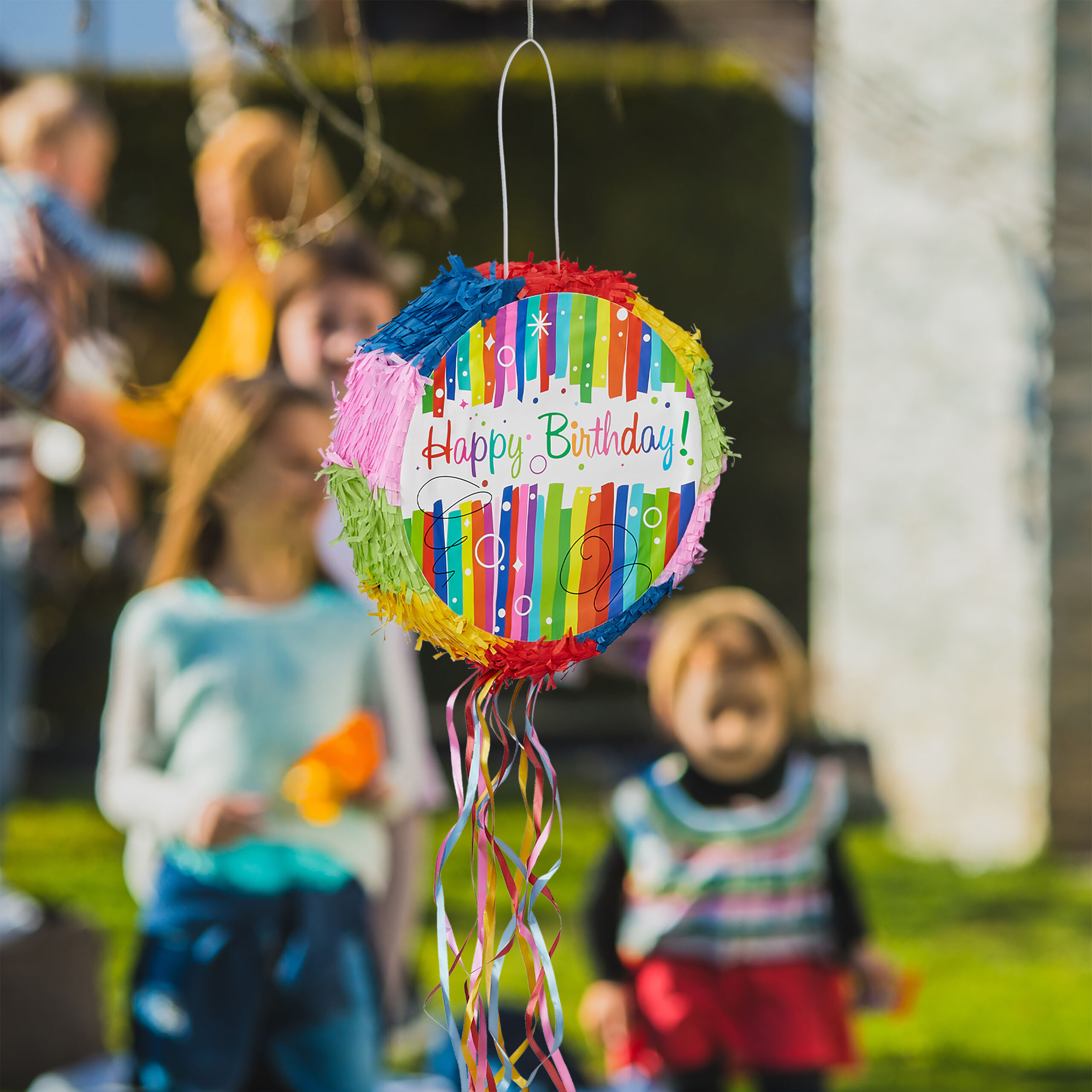 Eine bunte Happy Birthday Piñata hängt draußen, mit verschwommenen Kindern und grünem Gras im Hintergrund.