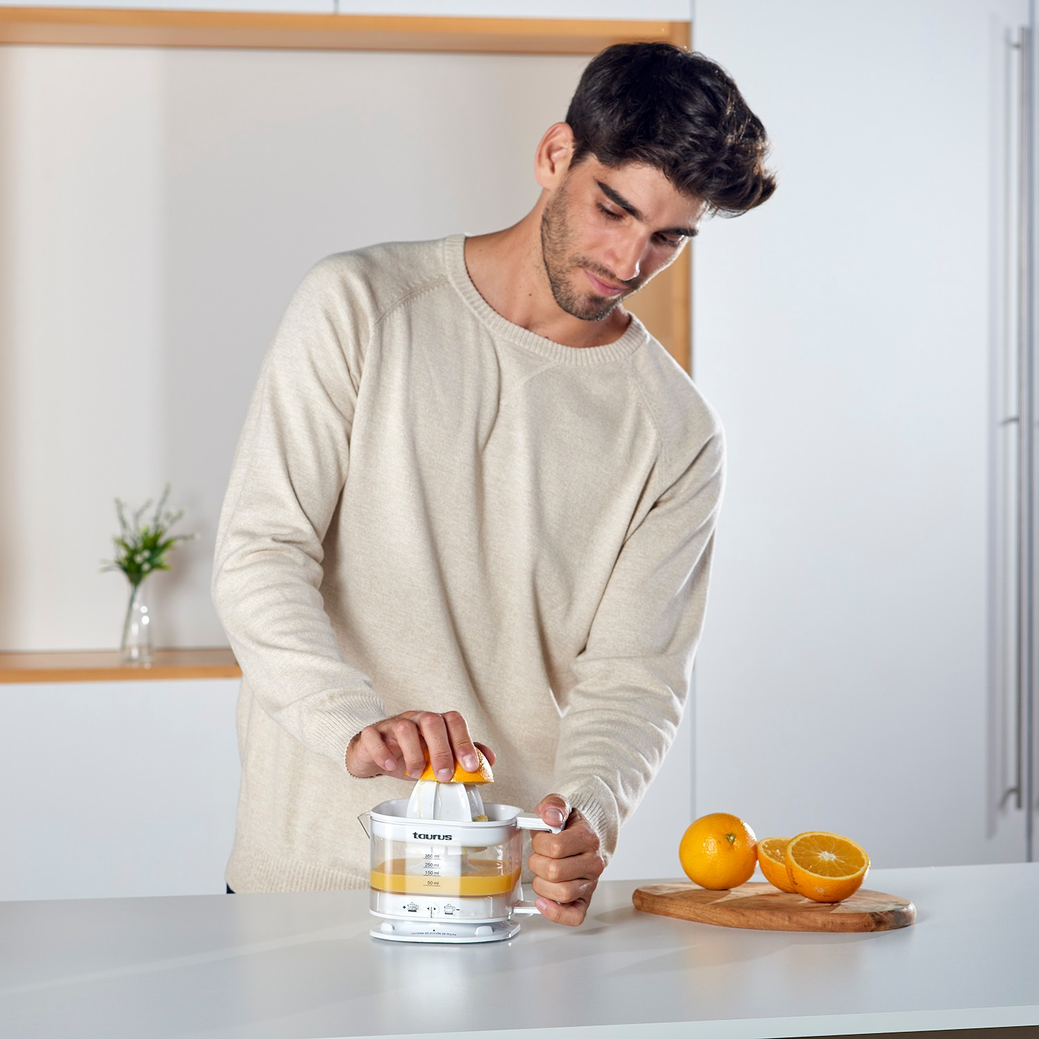 Un hombre con jersey beis exprime naranjas con un exprimidor eléctrico en una cocina.