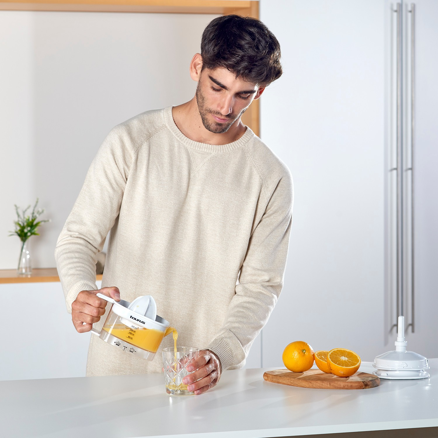Un hombre con jersey beis vierte zumo de naranja recién exprimido de un exprimidor en un vaso.