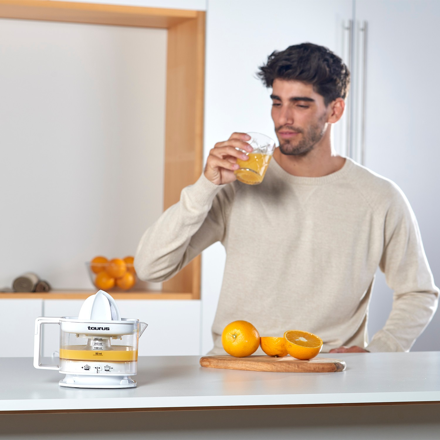 Un hombre bebe zumo de naranja en una cocina, con un exprimidor y naranjas frescas cerca.