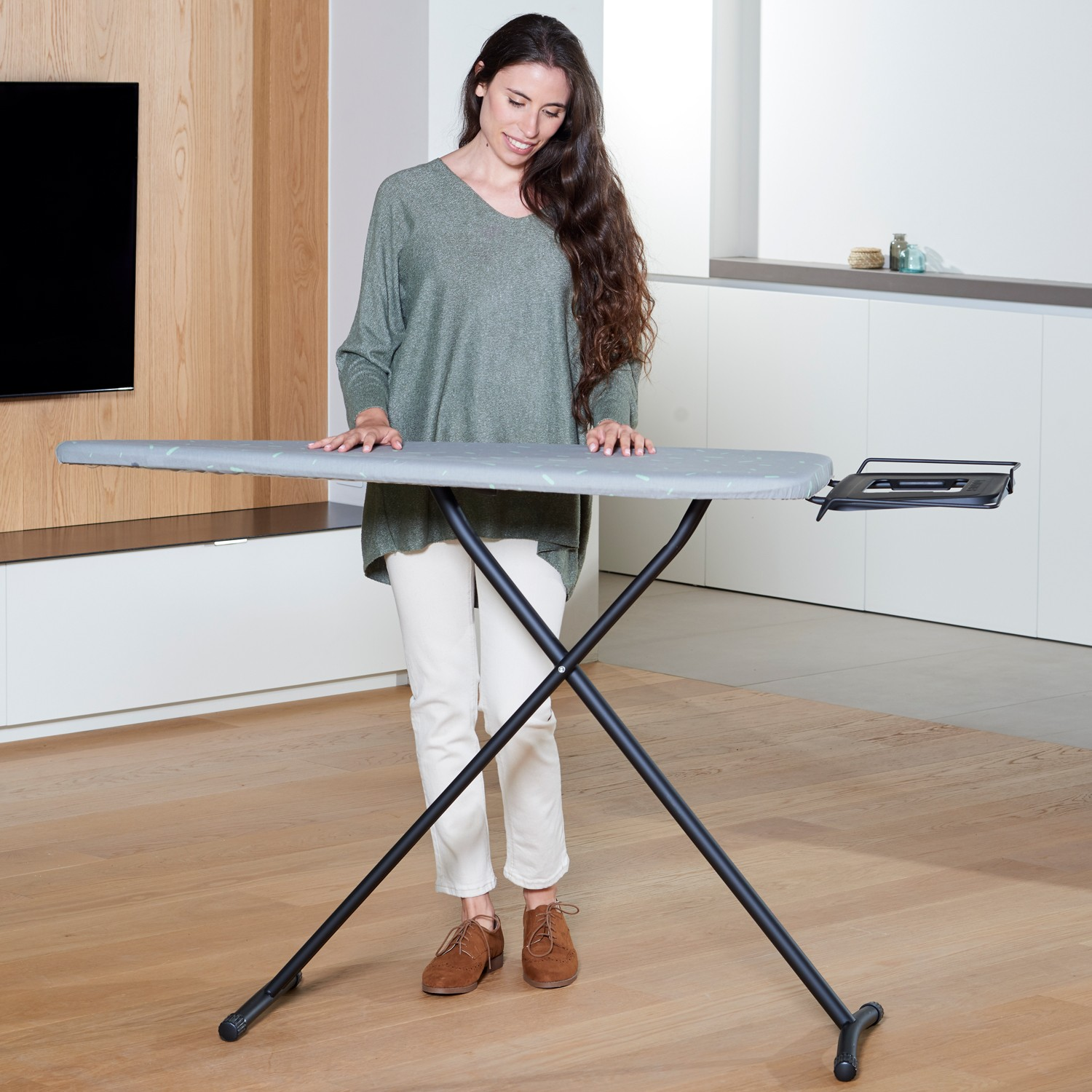 Una mujer con suéter verde está junto a una tabla de planchar moderna en una habitación luminosa.