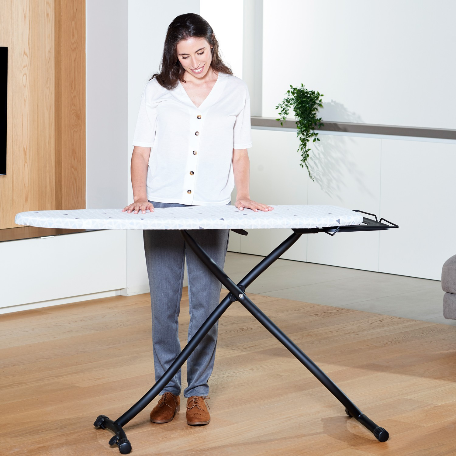 Una mujer con camisa blanca y pantalones grises está de pie junto a una tabla de planchar en una habitación luminosa.