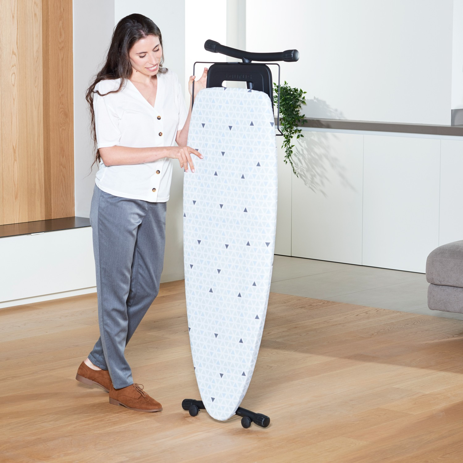 Una mujer con blusa blanca y pantalón gris está de pie junto a una tabla de planchar con patrón azul y blanco.