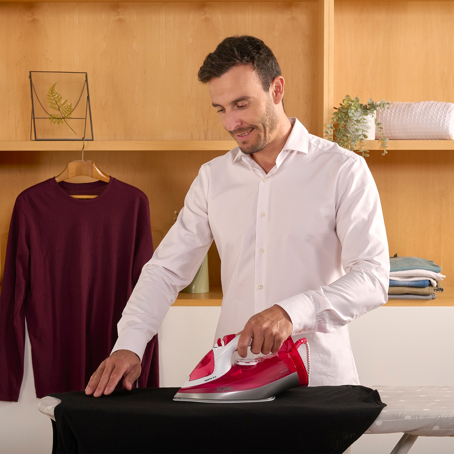 Un hombre con camisa blanca plancha una prenda negra con una plancha roja.
